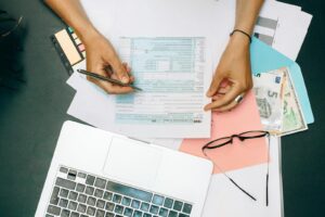 Mains écrivant sur des documents fiscaux avec un ordinateur portable, des lunettes et de la monnaie sur un bureau.